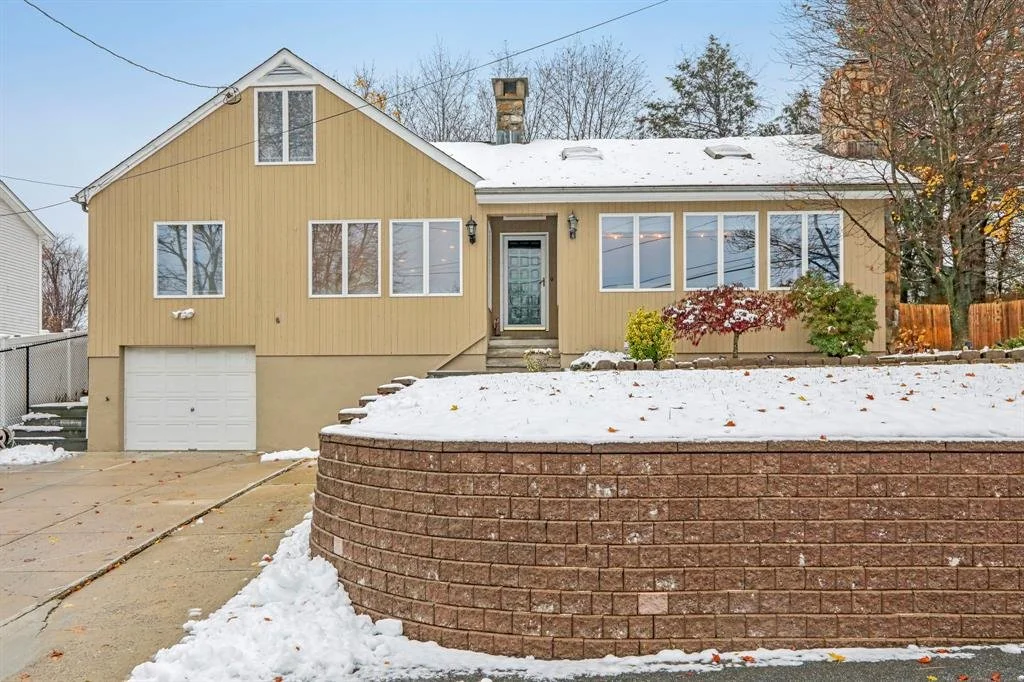 A house with a beige exterior, large front windows, and a garage, situated in a snowy yard with a visible front steps and a curved brick retaining wall.