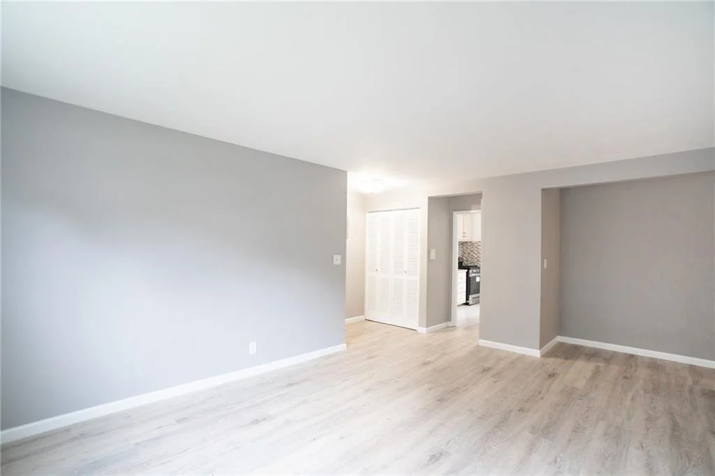 Empty living room with light gray walls, white baseboards, and light wood flooring, leading to a kitchen area with a stove and backsplash.