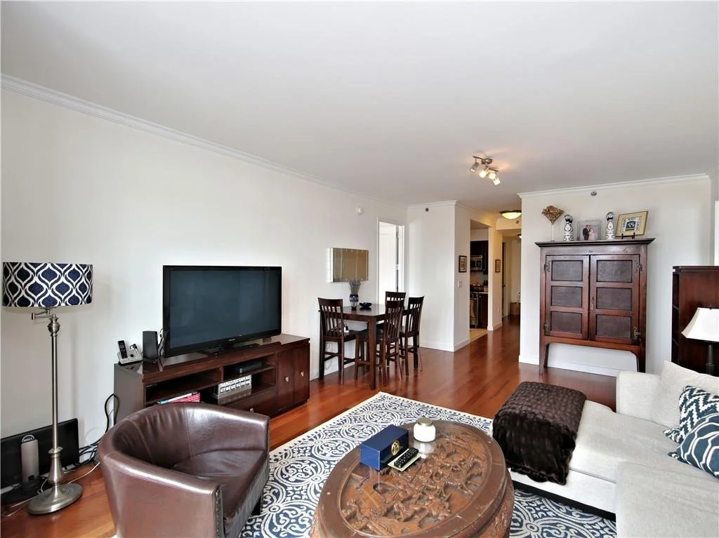 Living room with hardwood floors, white walls, a brown leather armchair, an upholstered sofa with pillows, a wooden coffee table, a flat-screen TV on a wooden stand, a tall standing lamp, a small dining table with four chairs, a large wooden armoire, and decorative items on top of the furniture.
