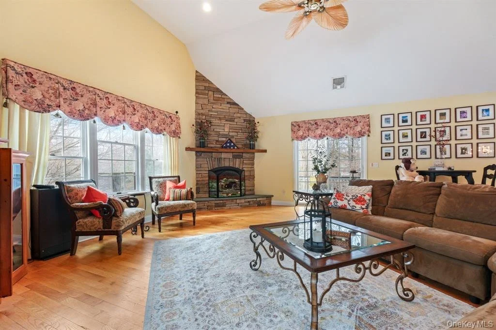 Living room with large windows, floral curtains, a brick fireplace, a brown sofa, vintage chairs, a glass-top coffee table, a light-colored area rug, and a gallery wall of framed photos.