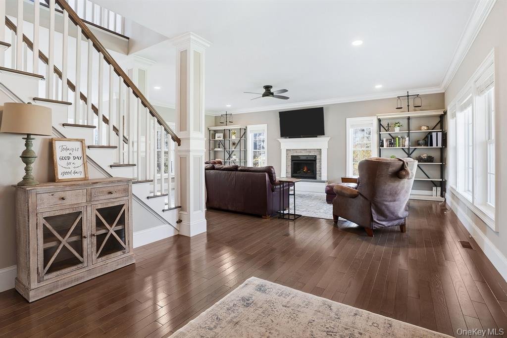 Living room with a staircase on the left side, a fireplace with a wall-mounted TV above it, and several chairs and a sofa arranged around the fireplace. There are large windows on the right side, a wooden floor, and a bookshelf on the right side.