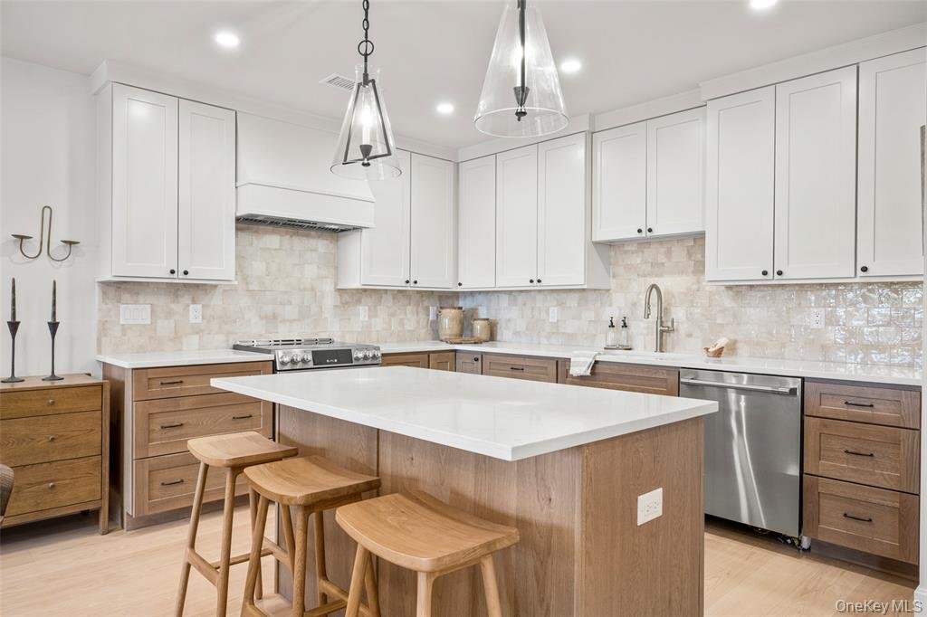 Modern kitchen with white upper cabinets, light wood lower cabinets, a kitchen island with a white countertop, three wooden bar stools, beige tiled backsplash, stainless steel dishwasher, and decorative items on the counters.