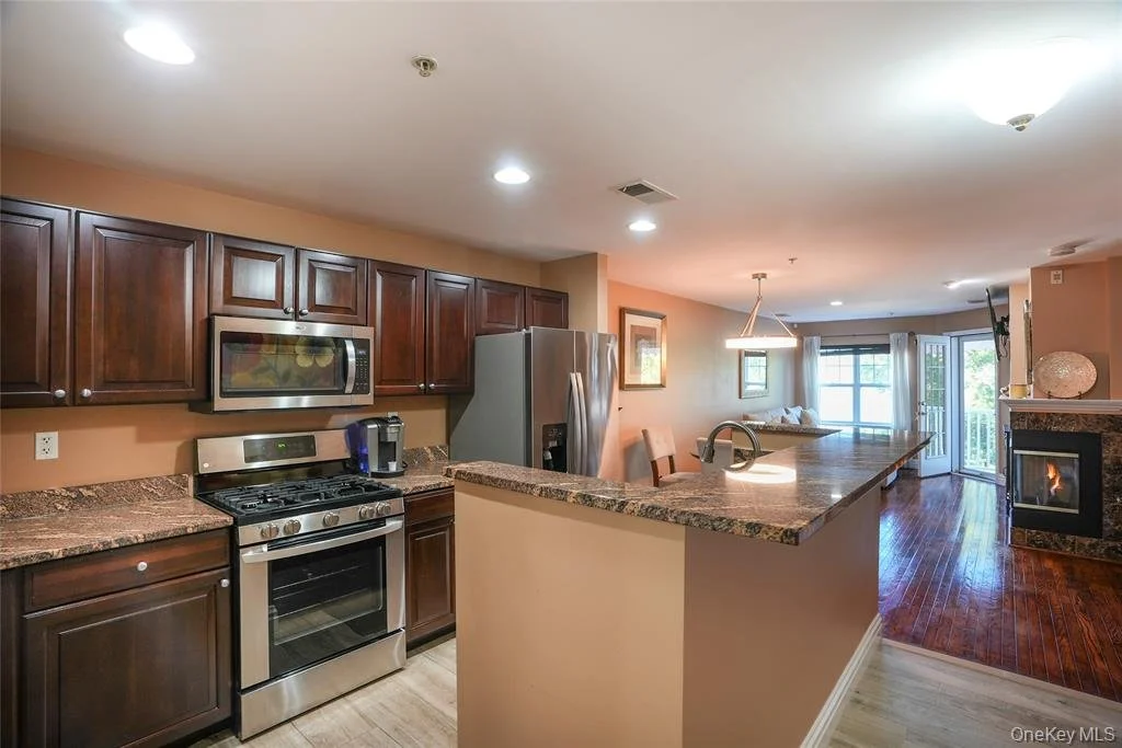 Kitchen with dark wood cabinets, stainless steel appliances including microwave, stove, and refrigerator, granite countertops, and an open living area with hardwood floors, a fireplace, and large windows.