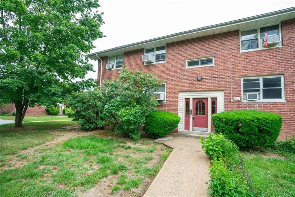 A red brick apartment building with a pink front door, six windows with air conditioning units, and greenery including bushes and trees.