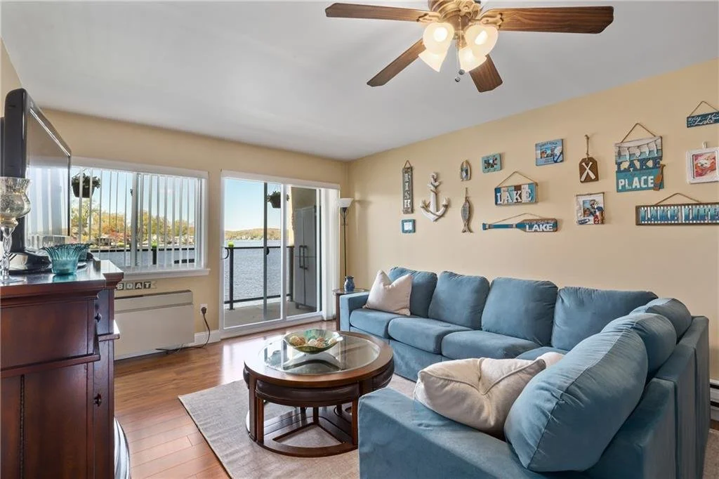 Living room with a large blue sectional sofa, wall art with lake and boat themes, a ceiling fan with lights, and sliding glass doors leading to a balcony with a lake view.