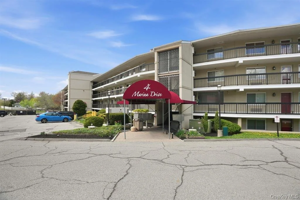 Multi-story apartment building with a red awning marked '4 Marina Drive' over the entrance, surrounded by landscaping and a parking lot with cars.
