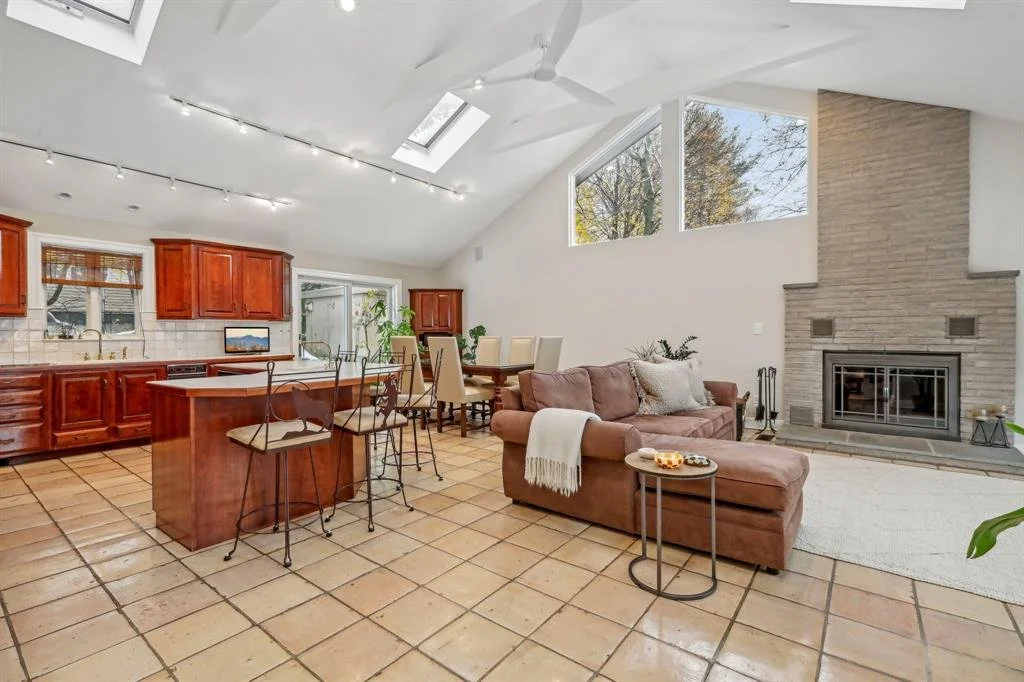 Open-concept living room and kitchen with tiled floor, brown cabinetry, and a fireplace, featuring high vaulted ceilings with skylights and large windows.