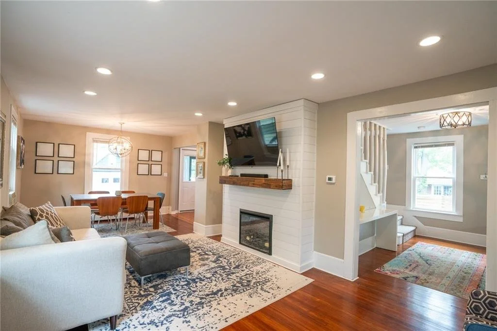 Living room with a white sofa, gray ottoman, fireplace, wall-mounted TV, and a dining area in the background.