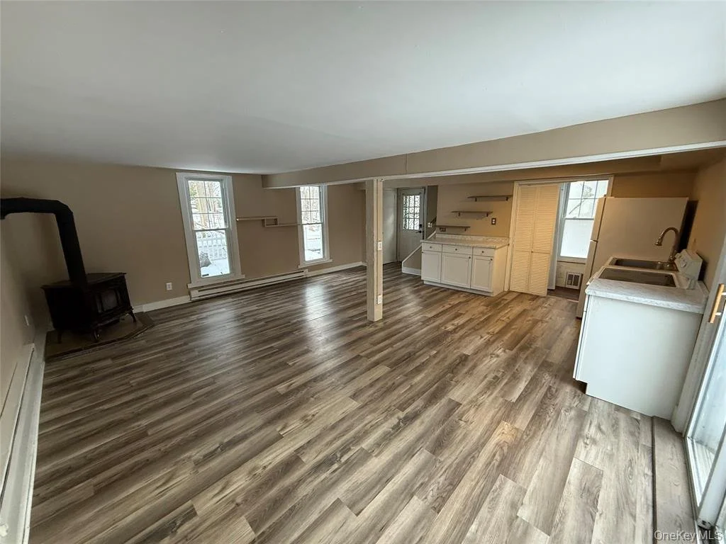 Empty living room with wood flooring, two windows, a small stove in the corner, built-in shelves and cabinets, a laundry area with sink, and a sliding glass door leading outside.