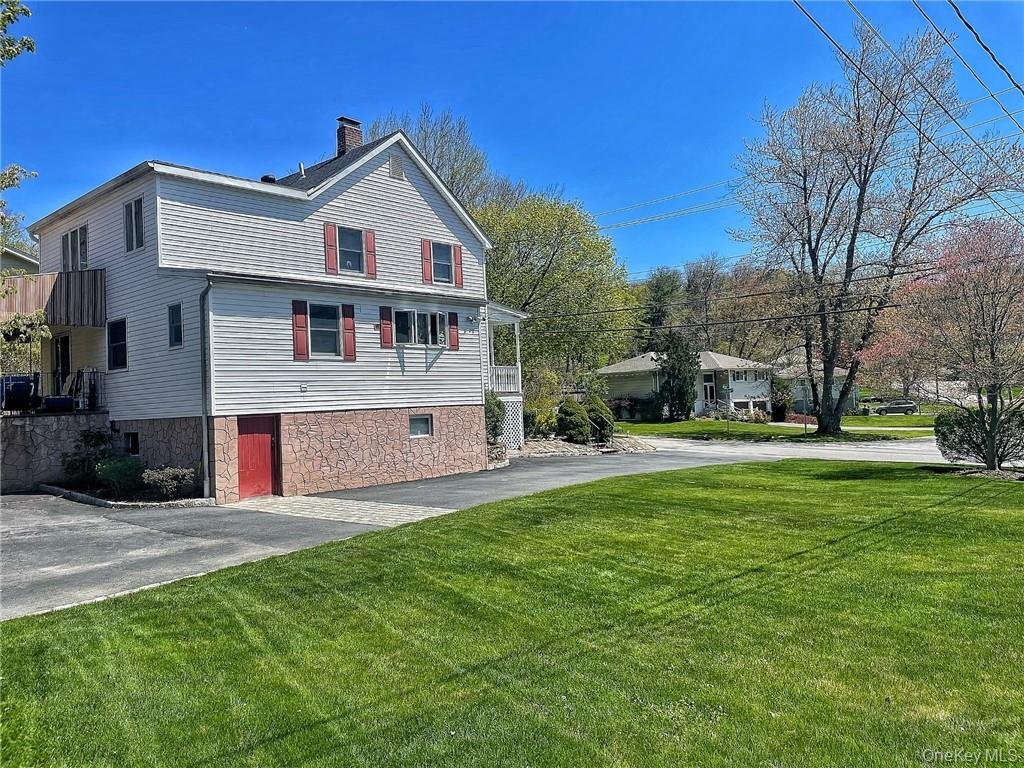 A two-story house with white siding and red shutters, a stone foundation, and a driveway in front. There is a well-maintained green lawn and trees in the background under a clear blue sky.