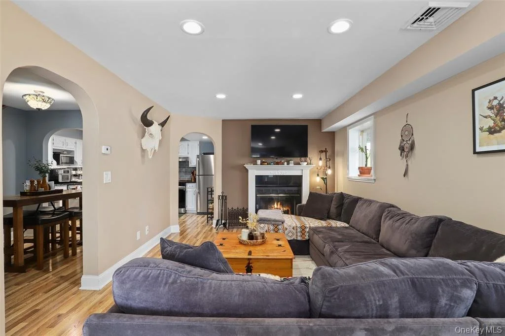 Living room with a gray sectional sofa, wooden coffee table, fireplace, TV above fireplace, wall decor including a dreamcatcher and framed picture, and a small window with a potted plant. An arched doorway leads to a dining area, and another door opens to a kitchen.