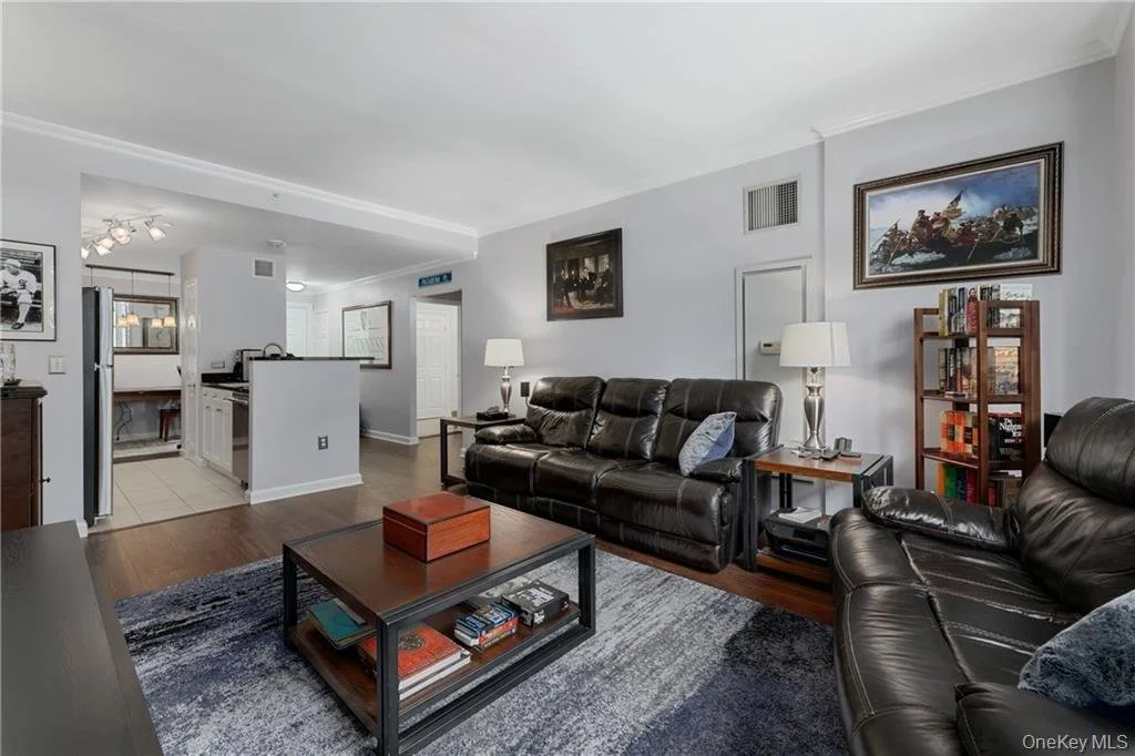 Living room with black leather sofas, wooden coffee table, side tables with lamps, a bookshelf, and framed artwork on the walls. Part of the kitchen and dining area are visible in the background.