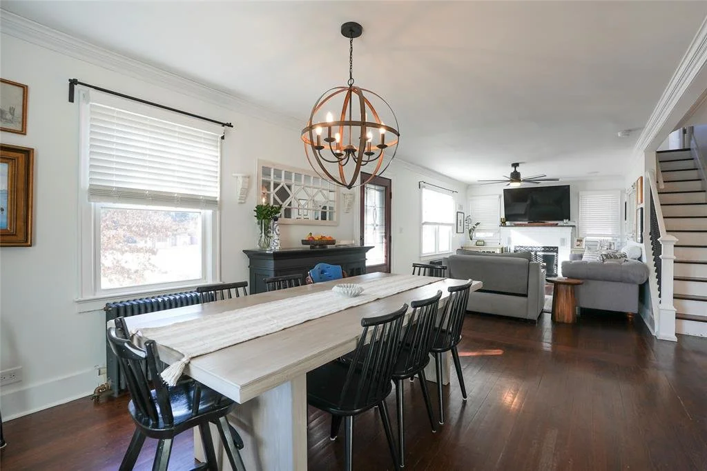 Open-concept dining and living room with wooden flooring, white walls, and large windows. The dining area has a rectangular table with six black chairs and a chandelier above. The living area includes two gray sofas, a mounted TV, a ceiling fan, and 