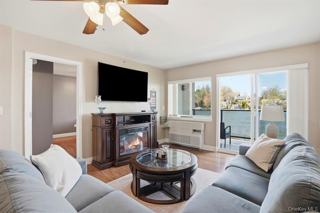 Living room with two light gray sofas, a wooden fireplace with a mounted TV, a round glass coffee table, a window with blinds, and a sliding glass door leading to a balcony with a view of the water.