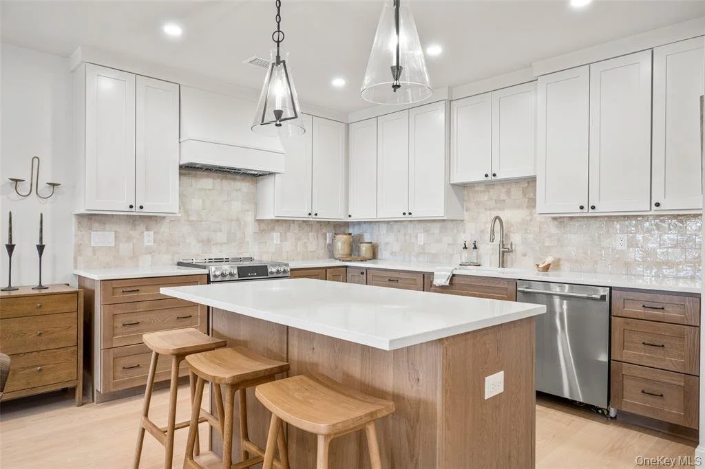 Modern kitchen with white upper cabinets, wooden lower cabinets, and a center island with a white countertop. There are two glass pendant lights hanging from the ceiling, a stainless steel dishwasher, and a beige tile backsplash. Wooden stools are pl