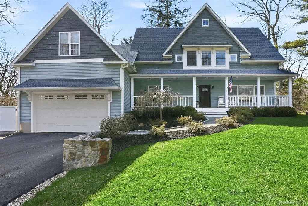 A large two-story house with light blue siding, white trim, and a dark roof. It features a front porch with white railings, a garage on the left, a well-maintained green lawn, and some shrubs and a small tree in the garden.