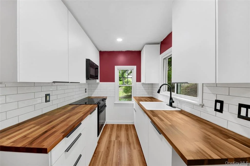 A modern small kitchen with white cabinets, wooden countertops, white subway tile backsplash, black appliances, including a microwave above the stove, and a window above the sink showing green outdoor scenery.
