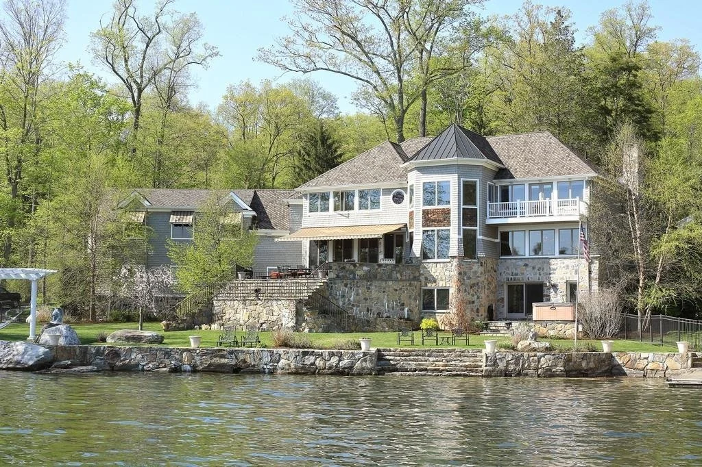 A large waterfront house with multiple levels, stone and siding exterior, and large windows, surrounded by trees.