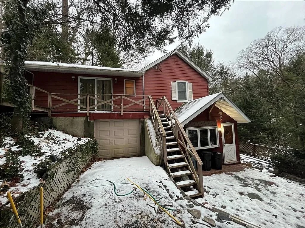 A two-story house with red siding, white window shutters, and snow-covered roof, surrounded by trees, with a wooden staircase leading from the upper deck to the snow-covered yard.