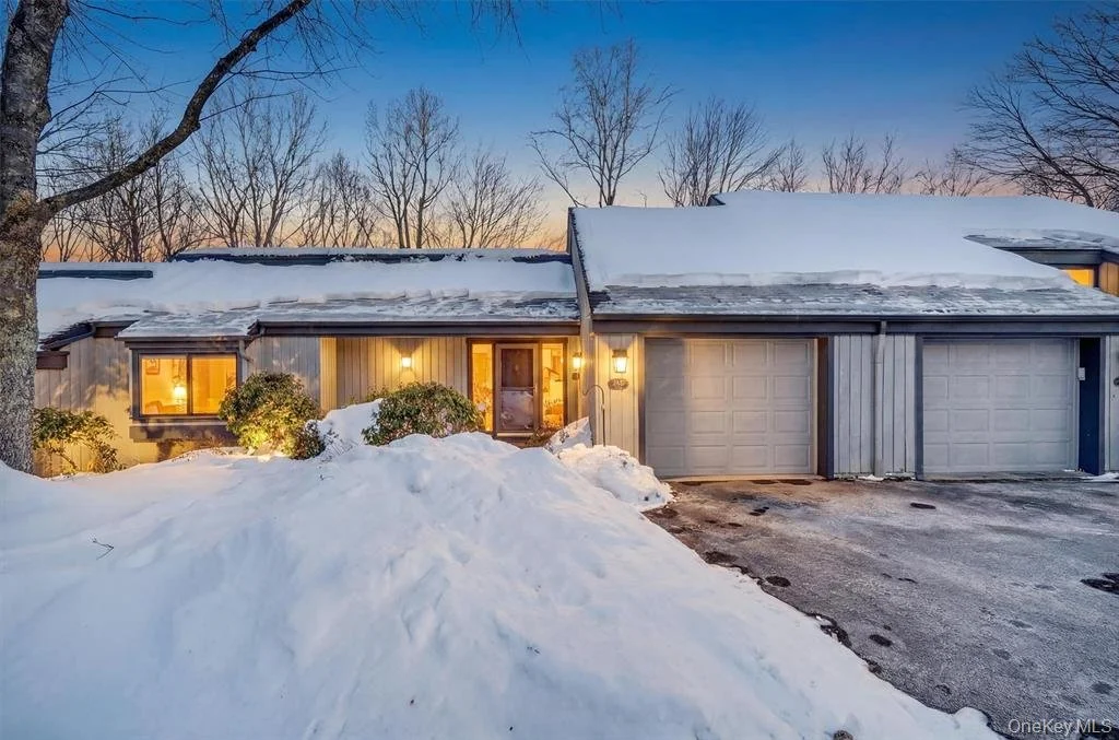 A snow-covered residential house at dusk with warm light coming from the windows, a large snow mound in the front yard, and a snow-covered roof and driveway.
