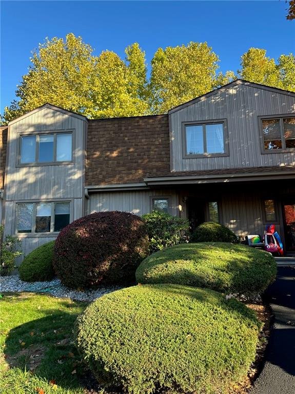 Front view of a two-story house with gray siding and brown roofing, surrounded by neatly trimmed bushes and trees with green foliage, under a clear blue sky.