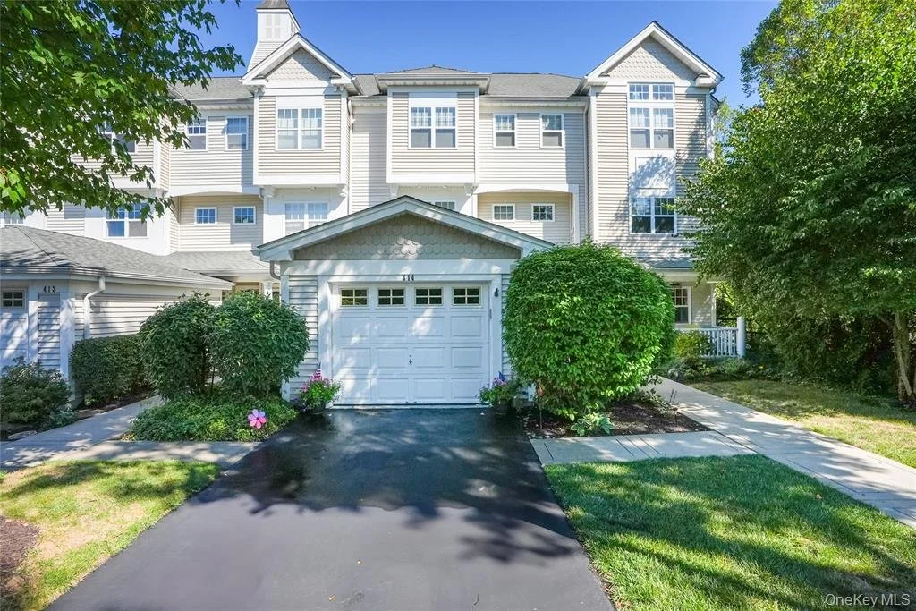 Front view of a multi-story townhouse with a white garage door, surrounded by green bushes and trees, with a paved walkway leading to the entrance.
