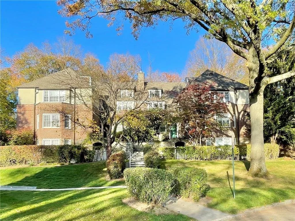 Multi-story residential building with a front yard, trees, and shrubs. Clear blue sky.