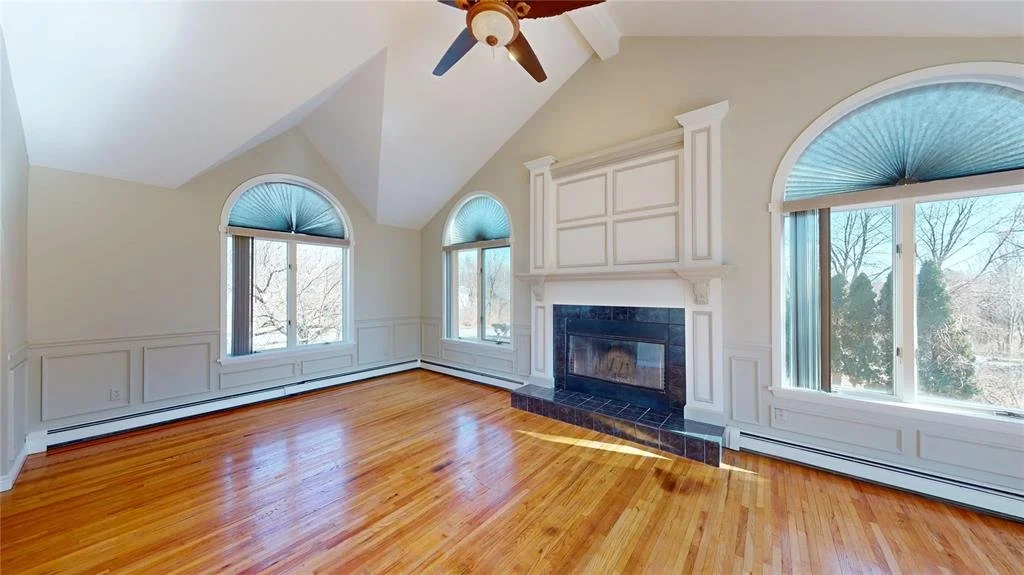 Empty living room with hardwood floors, large windows with blue shades, and a white fireplace with black tile accents.
