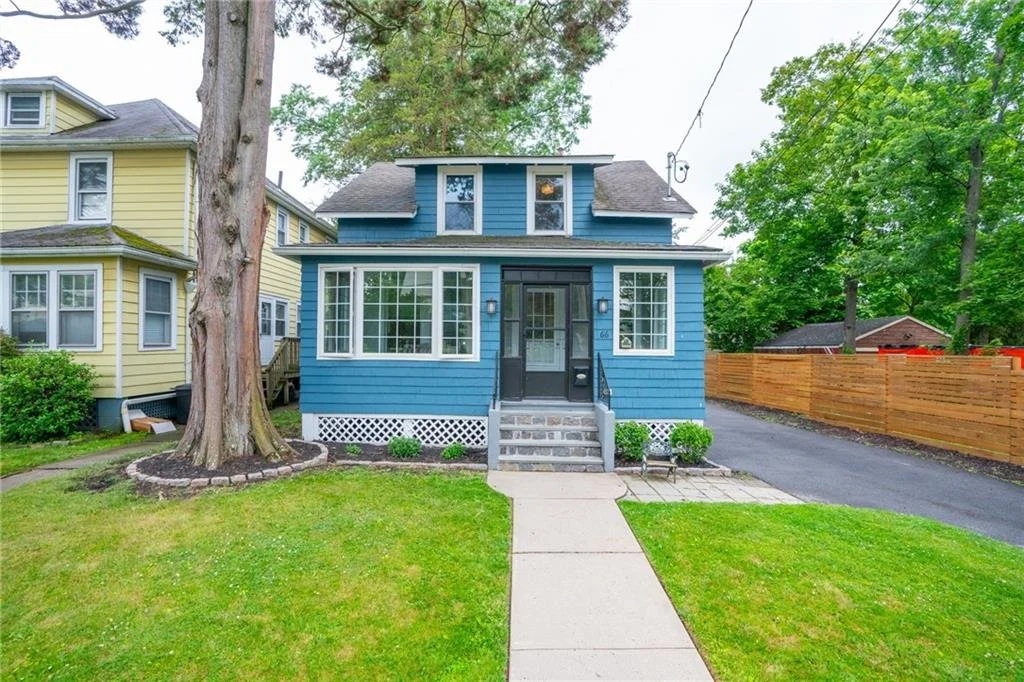 Blue two-story house with front steps, black door, surrounded by a green lawn, large trees, and a wooden fence to the right.