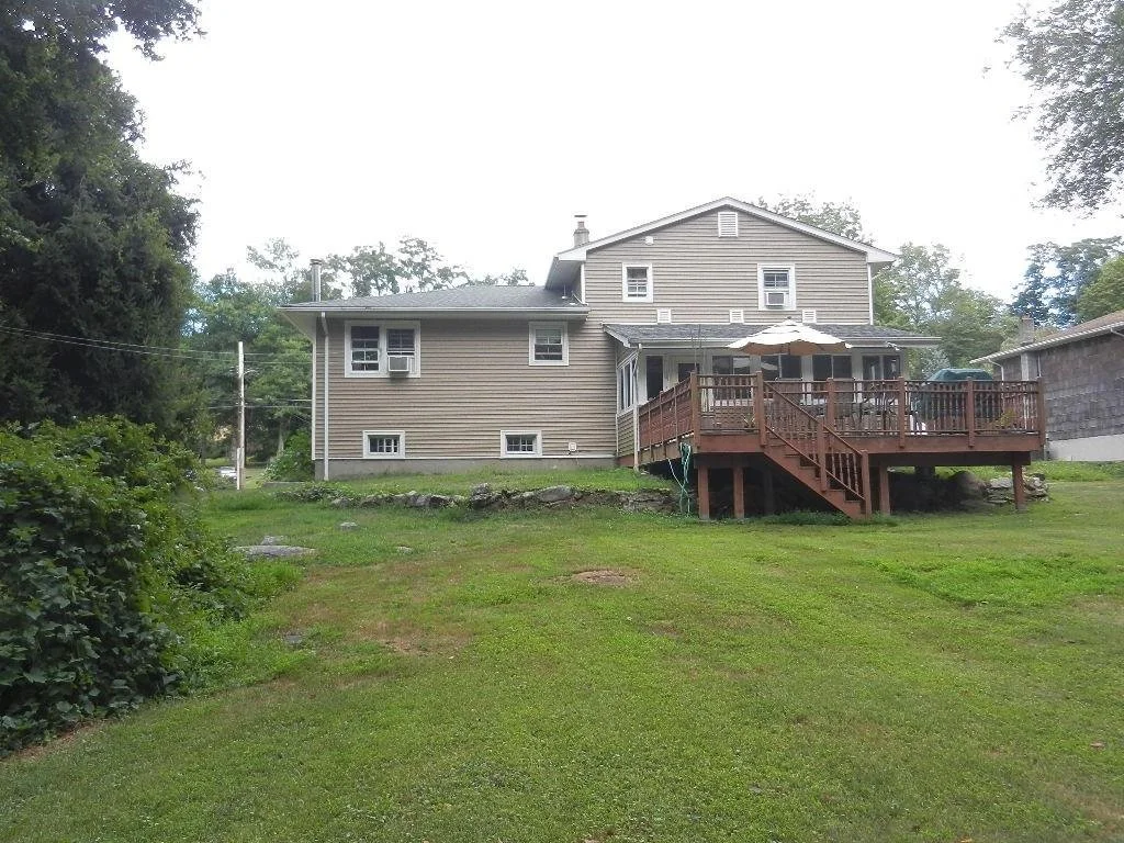 A two-story house with gray siding and a wooden deck in the backyard, surrounded by green grass and trees.