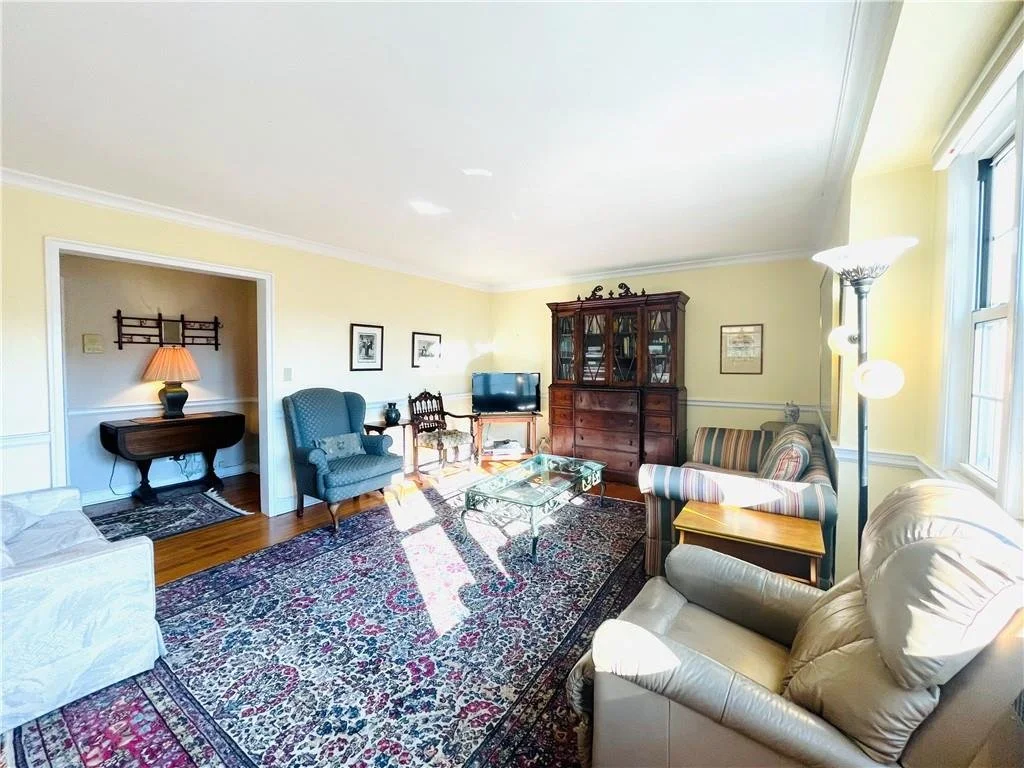 Living room with yellow walls, large window, beige and striped sofas, a blue armchair, a glass coffee table, a wooden china cabinet, and a patterned area rug.
