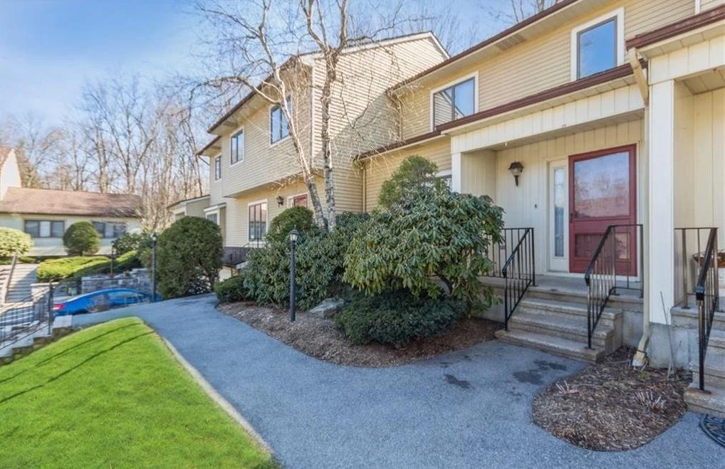 Exterior view of a beige townhouse with a red door, steps leading up to the entrance, and landscaped bushes and trees in front.