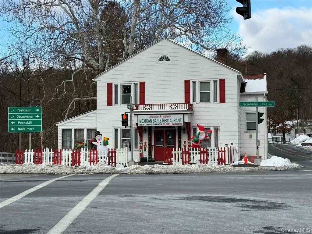 A white two-story building decorated for Christmas with inflatable snowman and Santa Claus, red fence, and Christmas and snow decorations, located at the corner of an intersection with street signs for Lake Peekskill, Lake Oscawana, and New York.