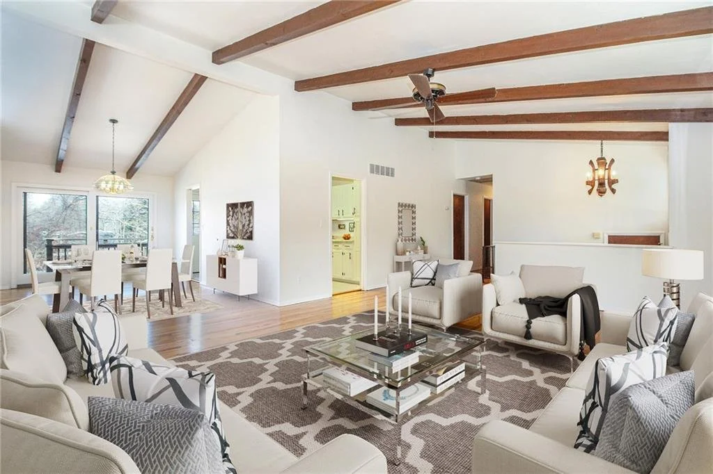 Bright open-concept living room and dining area with vaulted ceiling, wooden beams, white walls, and hardwood floors. Living room features white couches, decorative pillows, a glass coffee table with books and candles, a patterned rug, and a lamp. Di