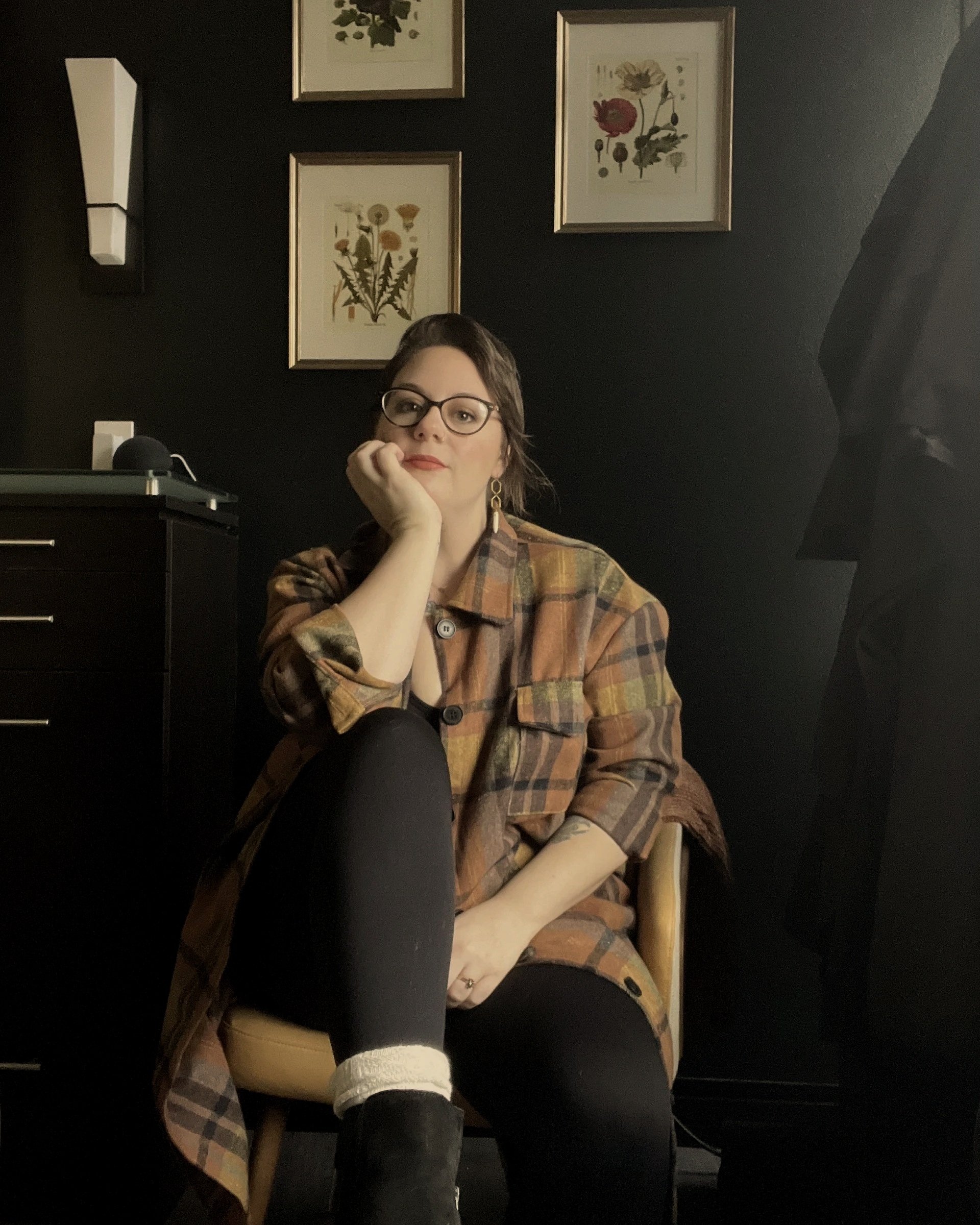 A woman with glasses and earrings sits at a table in a room with black walls. She is resting her head on her hand and looking at the camera. She wears a plaid shirt and black leggings with a white sock visible on her foot. Behind her, a black dresser with a device on top is visible. The wall is decorated with framed botanical prints.