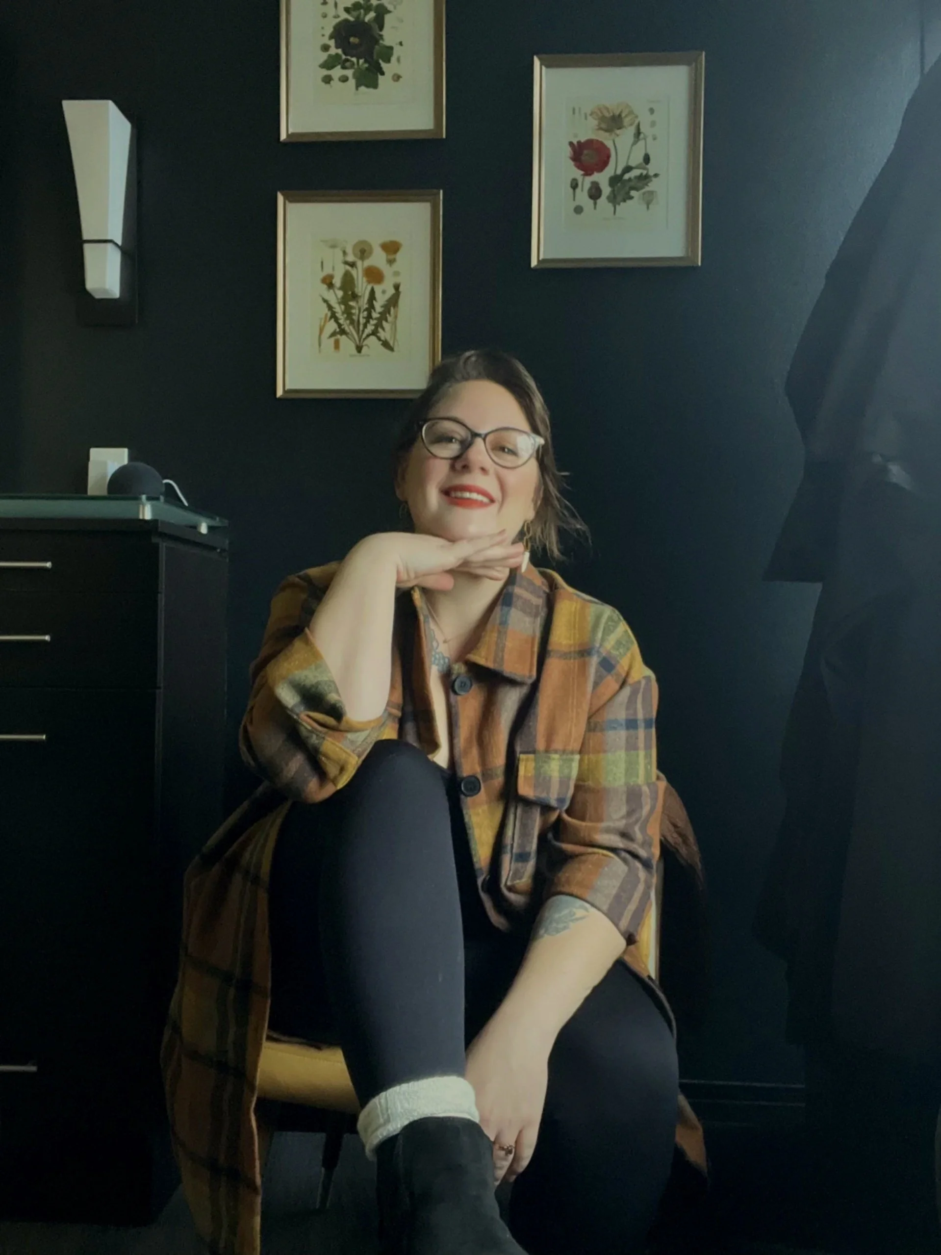 A woman with glasses and short hair, wearing a plaid jacket, sitting in a dark room with dark blue walls, smiling at the camera. Behind her are framed botanical prints on the wall.
