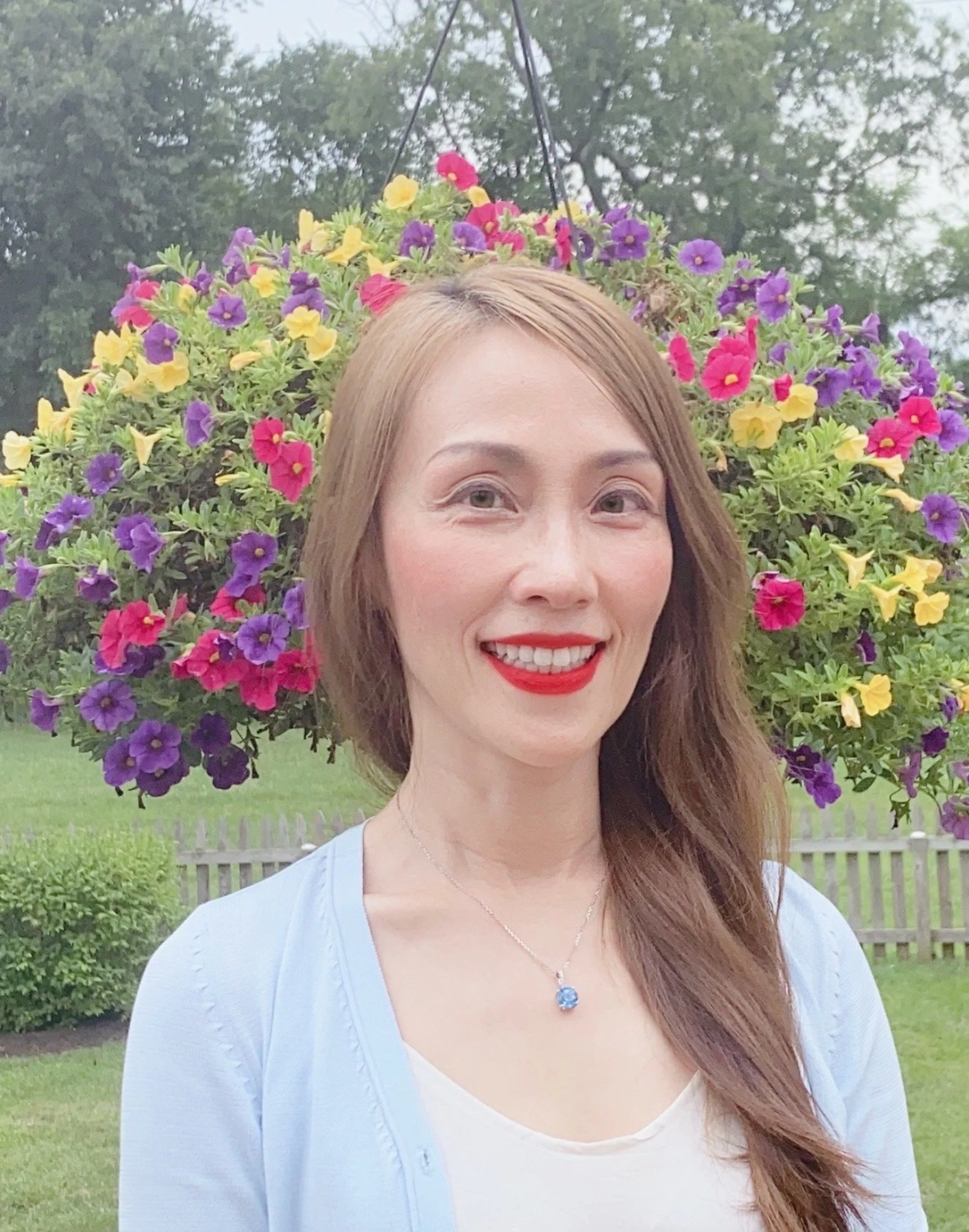 A woman with long, light brown hair and red lipstick smiling outdoors in front of a hanging basket of colorful petunias with purple, yellow, and pink flowers, a green lawn, trees, and a wooden fence in the background.