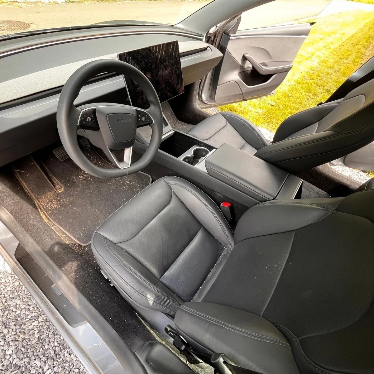 Interieur van een Tesla elektrische auto, met zwart stuur, groot touchscreen scherm, zwarte stoelen en centrale armleuning, parkeerplaats met grint buiten.