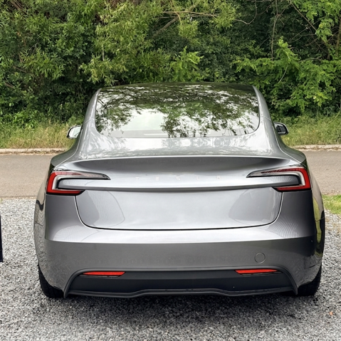 Achterkant van een zilverkleurige elektrische auto geparkeerd op een gravelweg, met groene bomen op de achtergrond.