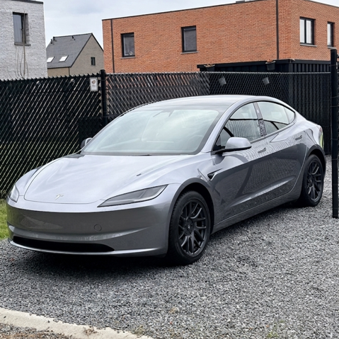 Zilveren Tesla Model 3 auto geparkeerd op een gravelterrein in een stad met huizen op de achtergrond.