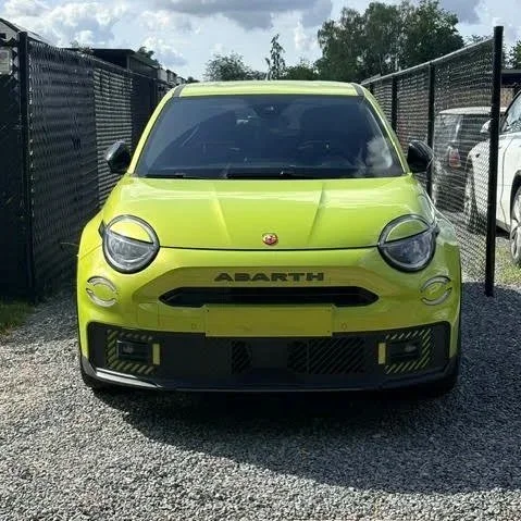 Acid Green Abarth 600e Scorpionissima met zwarte grille en sportbumper, geparkeerd tussen een metalen hek en andere auto's.