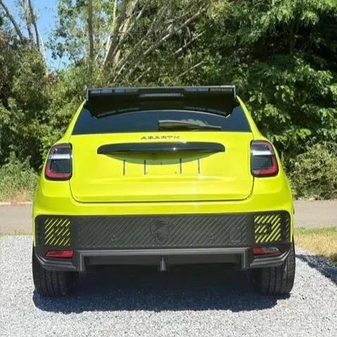 De achterkant van een felgele sportwagen met een zwarte spoiler en zwarte details, geparkeerd op een gravelweg met bomen op de achtergrond.