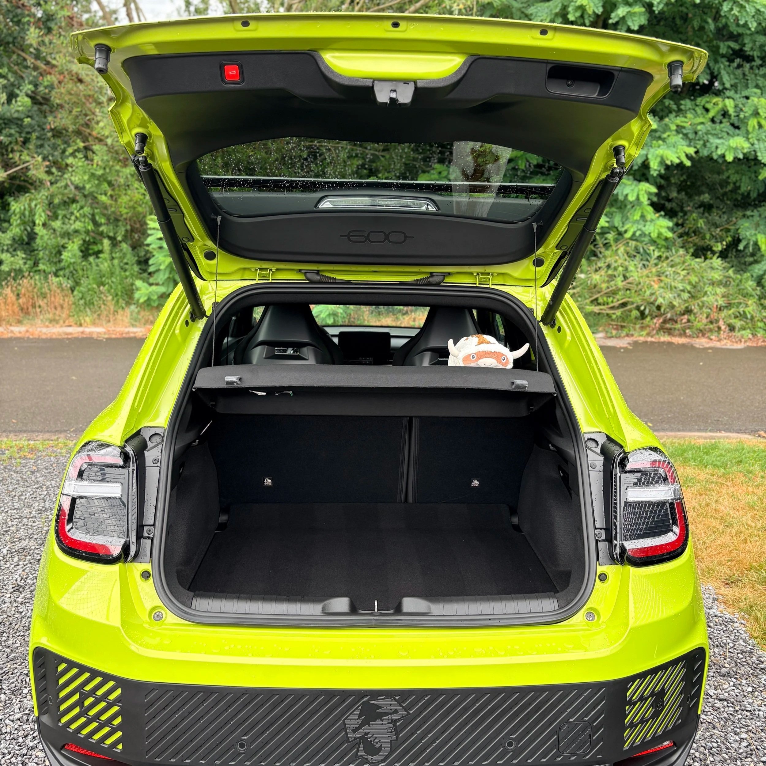 De achterklep van een felgele elektrische auto staat open, toont het lege bagageruimte met een zwart vloeroppervlak en een knuffel in de auto. Bomen en grasperken zijn op de achtergrond zichtbaar.