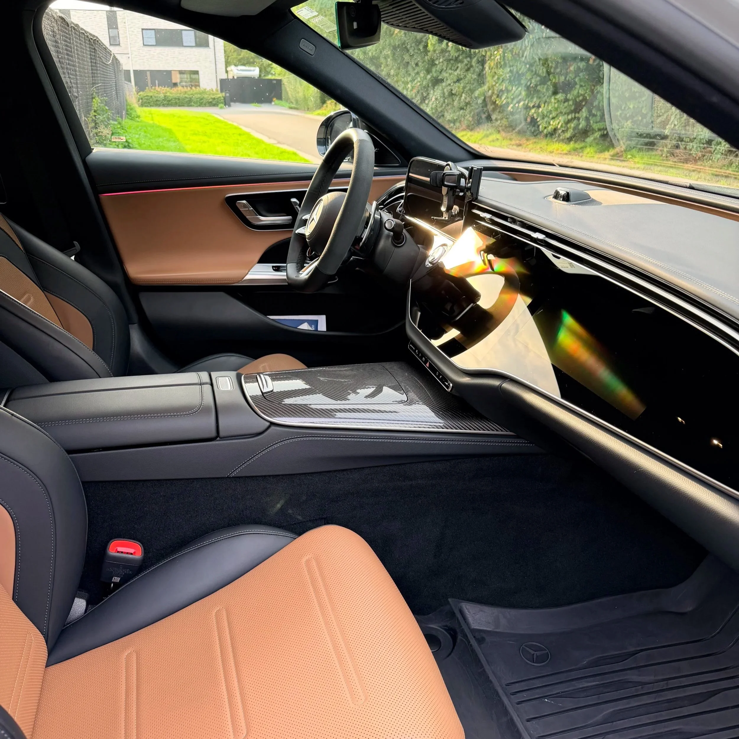 Interieur van een luxe auto, met oranje leren stoel, stuur en dashboard met een groot scherm, uitzicht op een straat met groen gras en een modern gebouw.