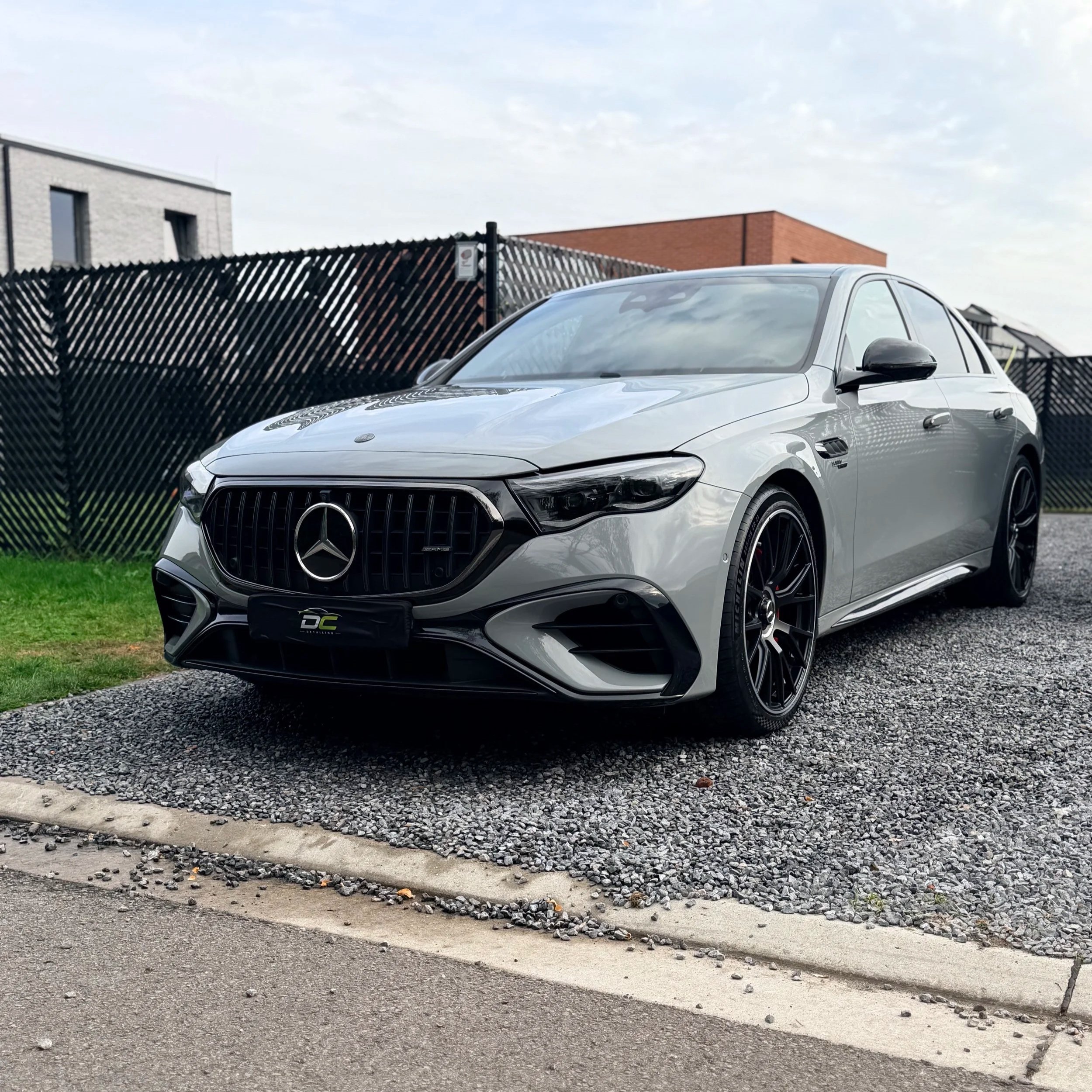 Een zilveren Mercedes E53 AMG naast een grasveld en een metalen hek, met moderne gebouwen op de achtergrond onder een bewolkte hemel.