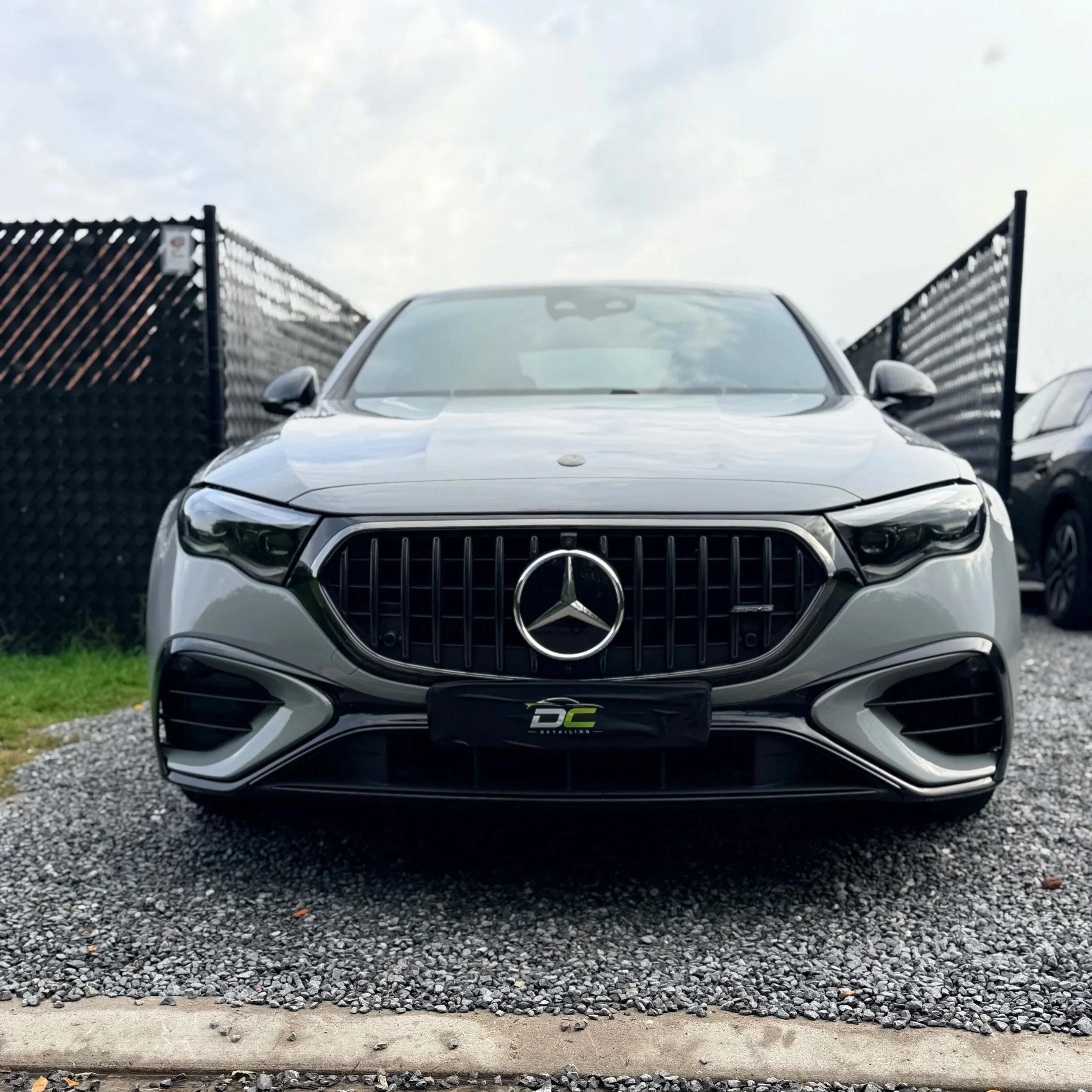 Een zilveren Mercedes AMG E53 geparkeerd tussen twee zwarte hekken met gaten, op een plaats met grind en een betonnen rand. Het weer is bewolkt.