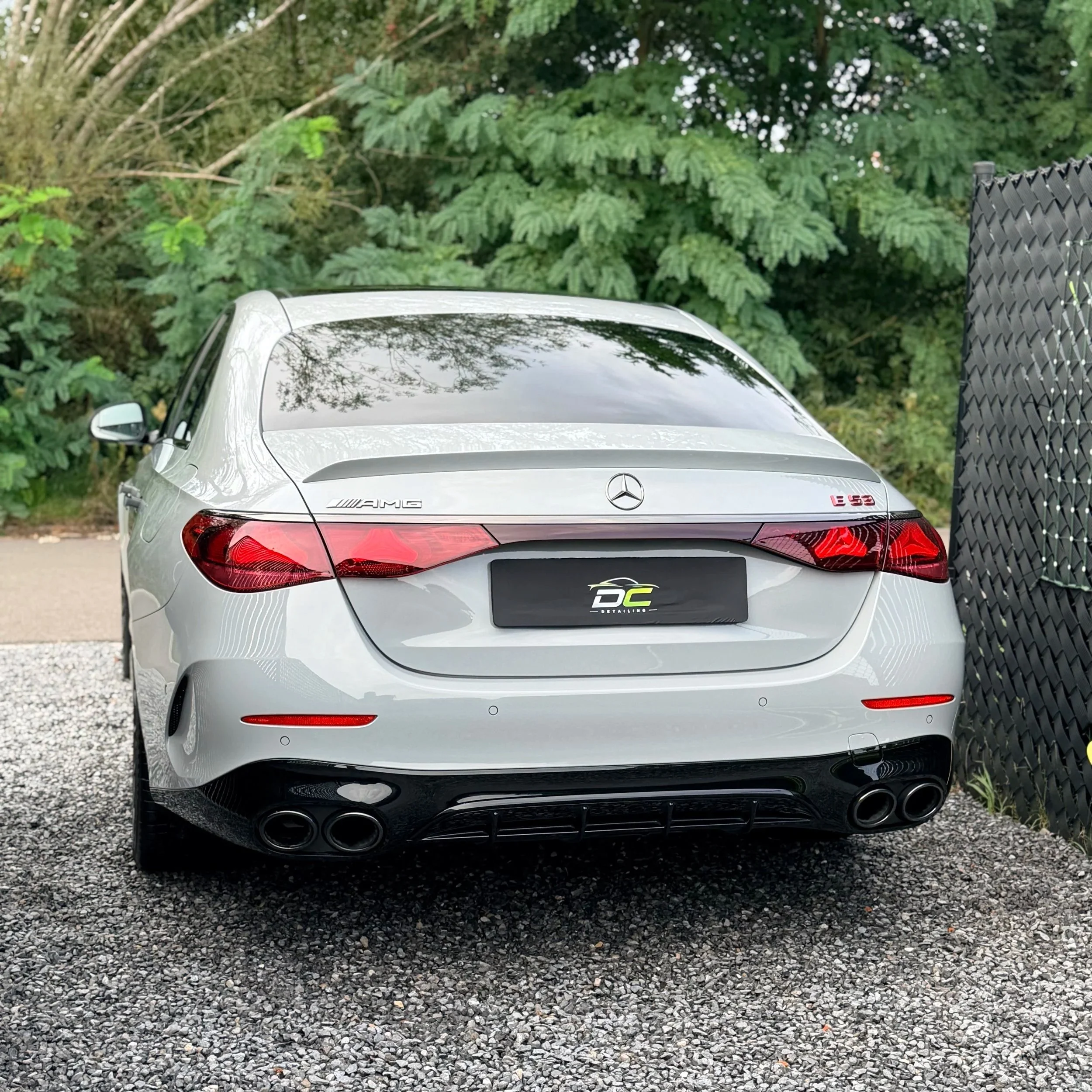 De achterkant van een zilveren Mercedes-Benz auto geparkeerd op een gravelweg, met groene bomen op de achtergrond en een vertikaal zwart hek aan de rechterkant.
