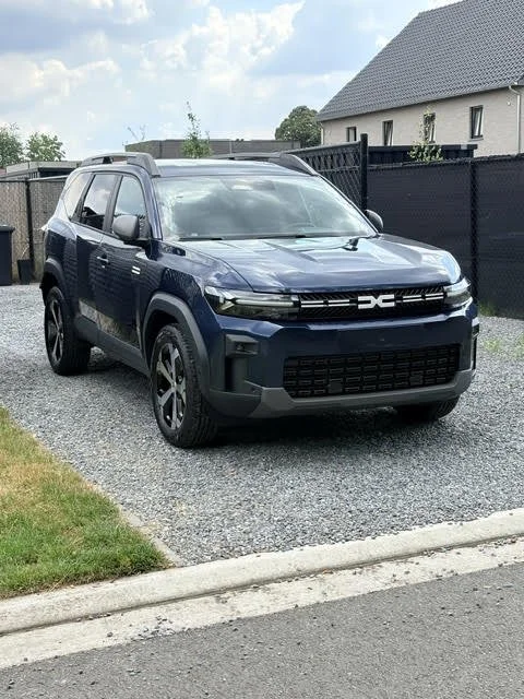 Schuin aanzicht van een donkerblauwe Dacia Bigster Hybrid SUV geparkeerd op een oprit van grind met op de achtergrond een modern hekwerk en huis