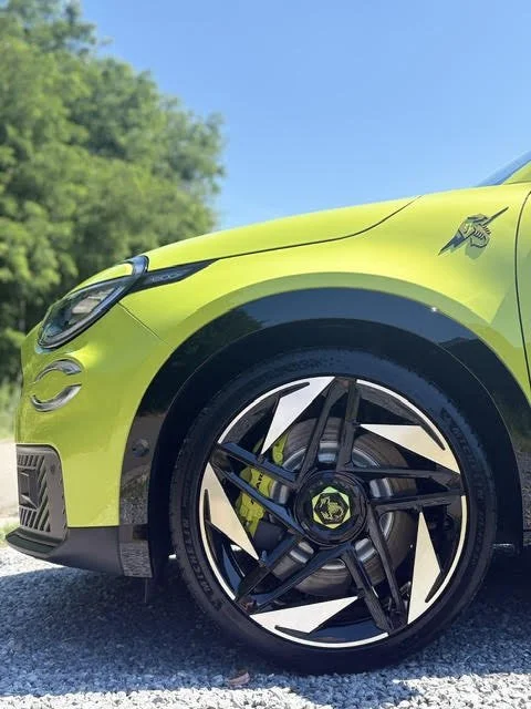 De zijkant van een Acid Green Abarth 600e Scorpionissima met zwarte en zilveren aerodynamische velg, geparkeerd in een groene, bosrijke omgeving