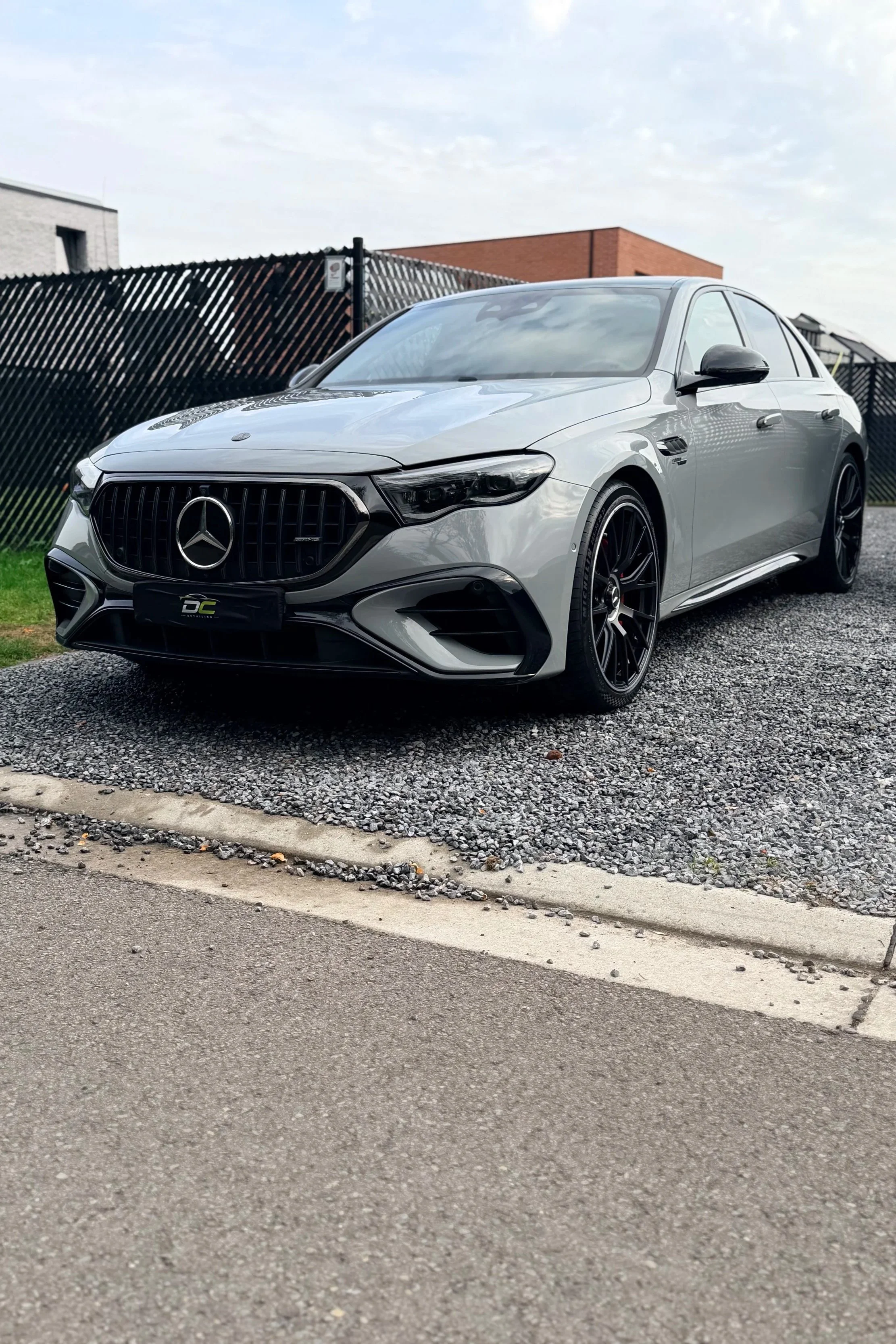 Een zilverkleurige Mercedes-Benz AMG auto geparkeerd op een oprit met enkele gebouwen en een hek op de achtergrond.